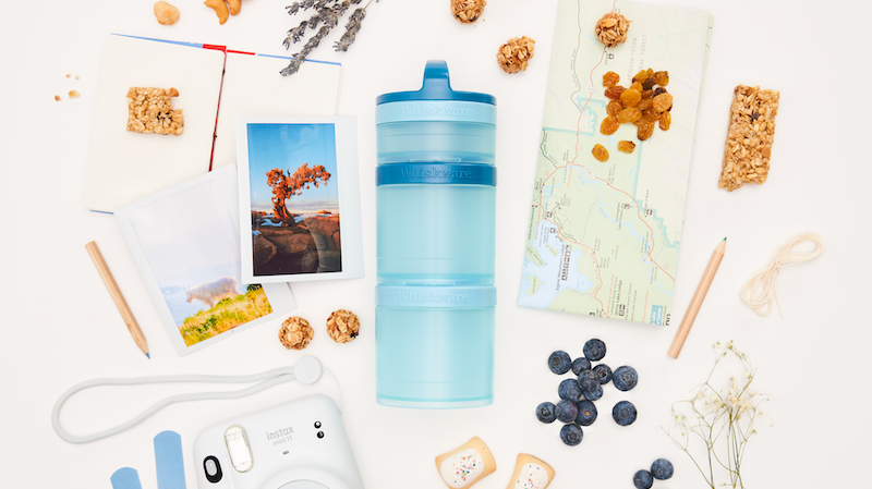 A light blue Whiskware snacking container surrounded by travel items like a map, photos, and a camera, plus snacks including blueberries, granola bites, and cashews on a white background.