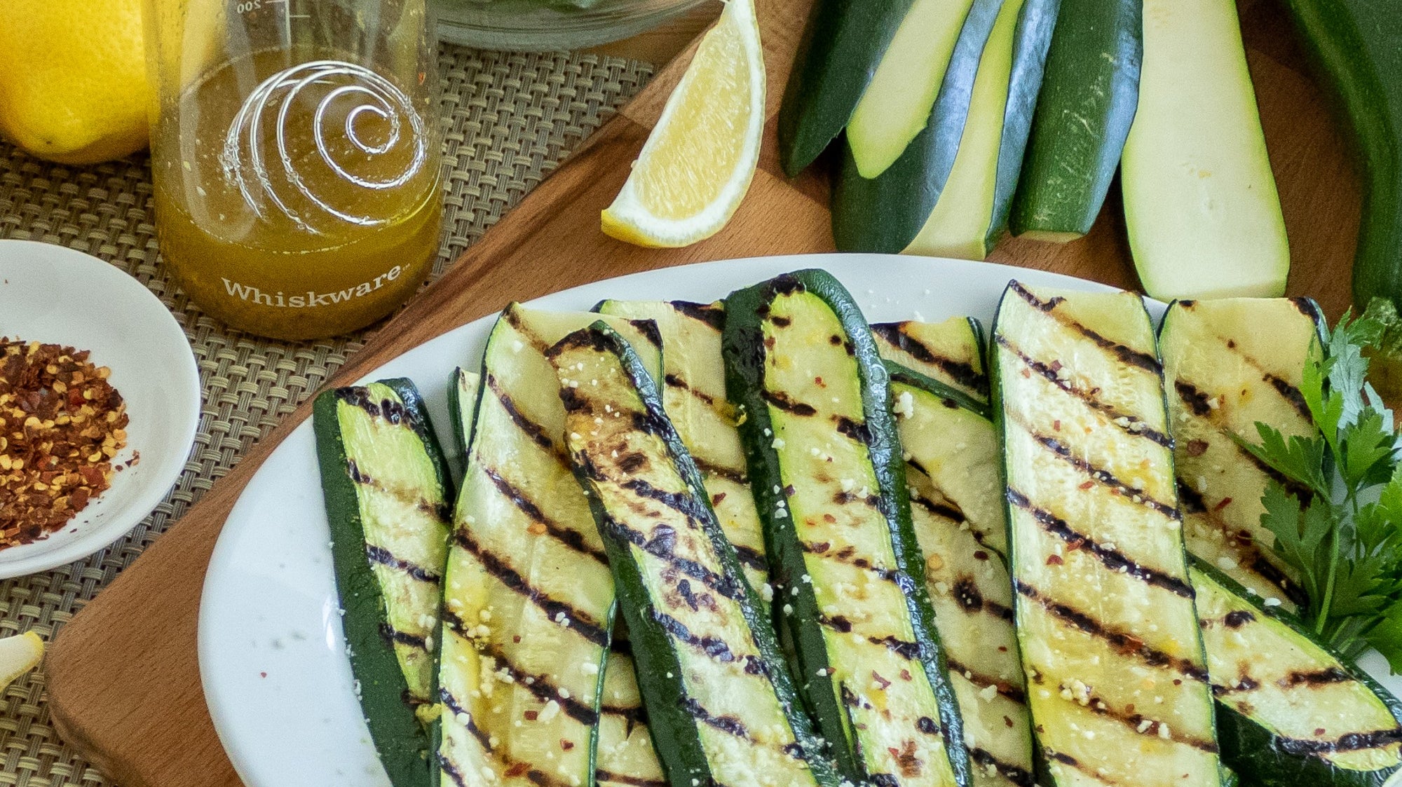 Grilled Zucchini cut in half with visible grill marks. behind them are uncooked zucchini, a Whiskware Dressing Shaker, a lemon, and a bowl of salad greens.