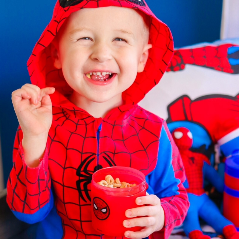 A little boy holding a spiderman snacking container while wearing a spiderman costume and smiling.