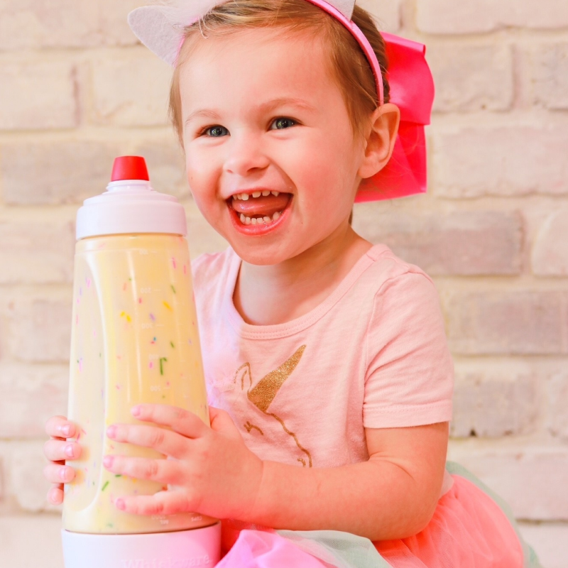 A little girl in a unicorn costume holding a batter mixer filled with funfetti batter.