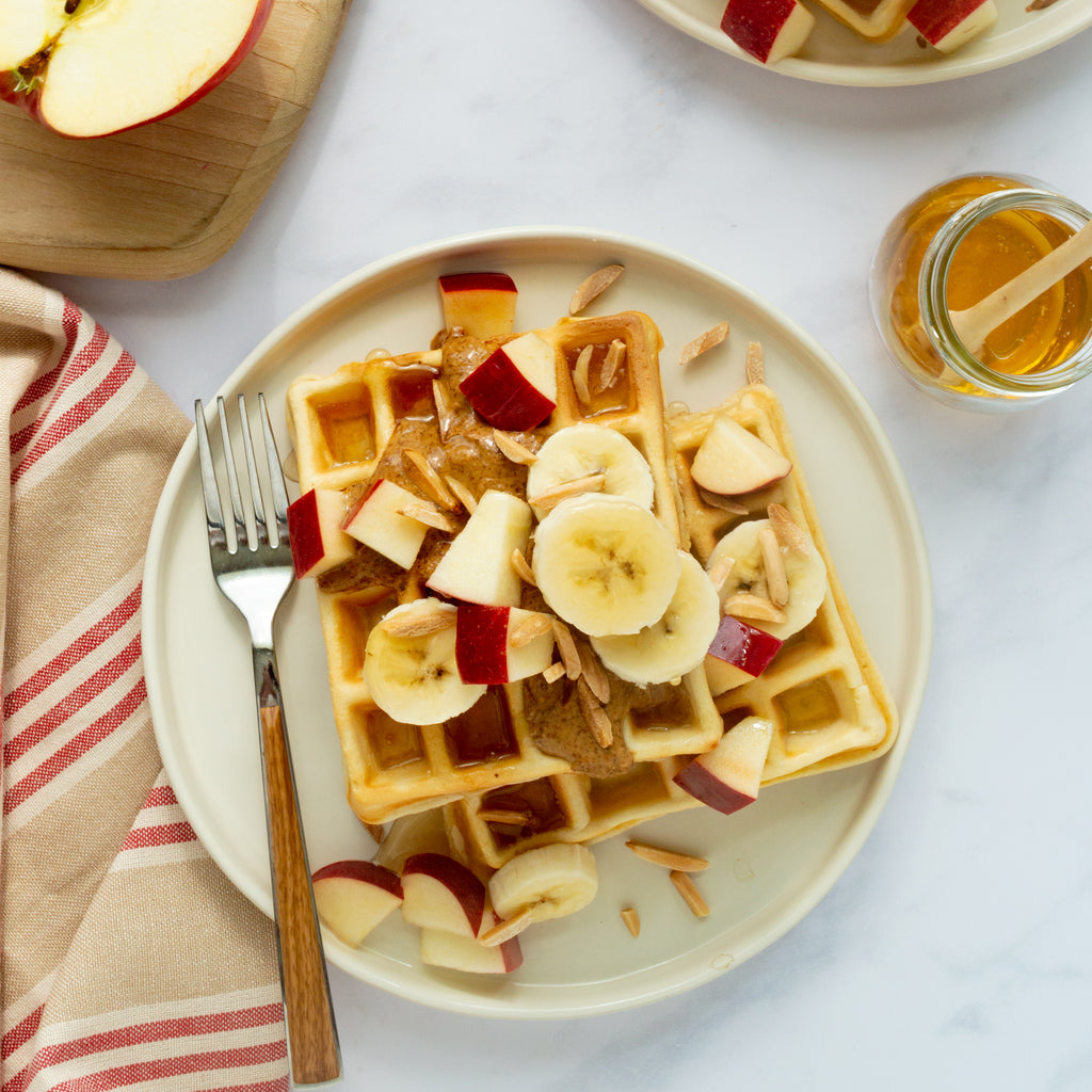 a plate of waffles stacked two high garnished with banana slices, apple chunks, and walnuts. Surrounding the plate is half of an apple, a jar of honey, and a red striped, tan napkin.