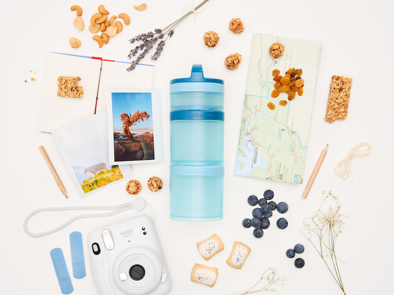 A light blue Whiskware snacking container surrounded by travel items like a map, photos, and a camera, plus snacks including blueberries, granola bites, and cashews on a white background.