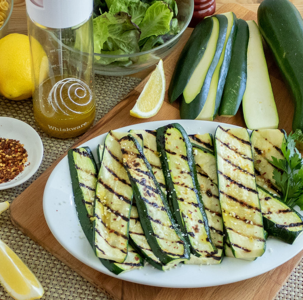 Grilled Zucchini cut in half with visible grill marks. behind them are uncooked zucchini, a Whiskware Dressing Shaker, a lemon, and a bowl of salad greens.