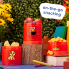 Red snack containers with cartoon characters on a blue surface with flowers and books in the background, labeled 'on-the-go snacking'.