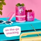 Pink snack containers with Barbie logo on a blue surface with sunglasses and berries, featuring the text 'on-the-go snacking'.