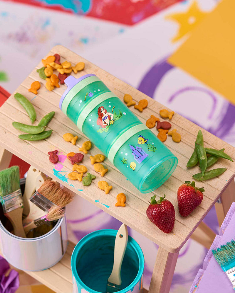 Children's play area with a green snack container featuring an Ariel design on a wooden stool with snacks & paint supplies.