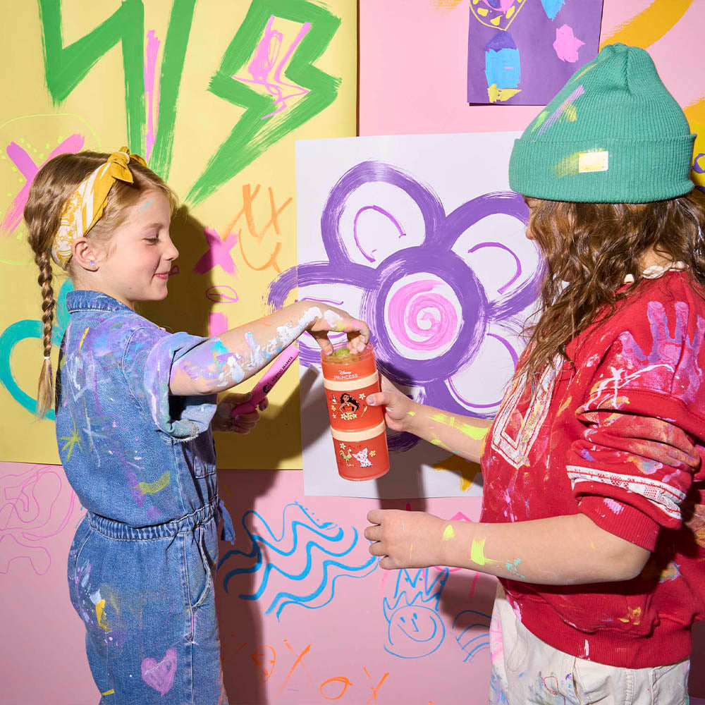 Two children sharing snacks from a Moana container with children's artwork hanging on the wall behind them.