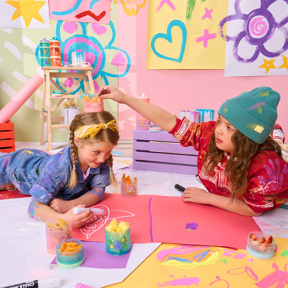 Two children playing with art supplies and snacking from Disney Princess Storybook containers in a colorful room.