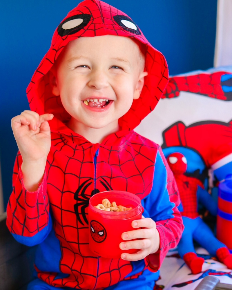 A little boy holding a spiderman snacking container while wearing a spiderman costume and smiling.