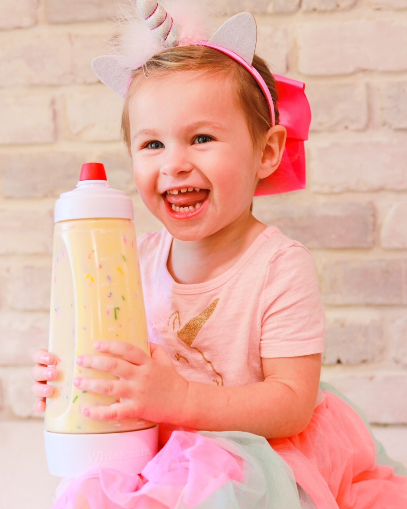 A little girl in a unicorn costume holding a batter mixer filled with funfetti batter.