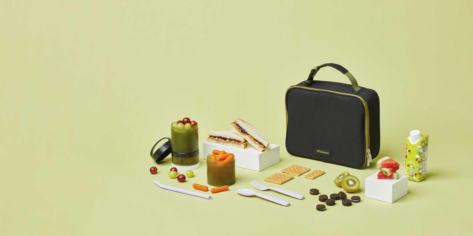 Whiskware Lunchbox and Snacking Container displayed on a green background with various snacks and utensils.