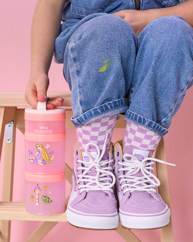 Girl wearing purple high-top sneakers holding a snack container with a Rapunzel Disney design, sitting on a stool against a pink background.